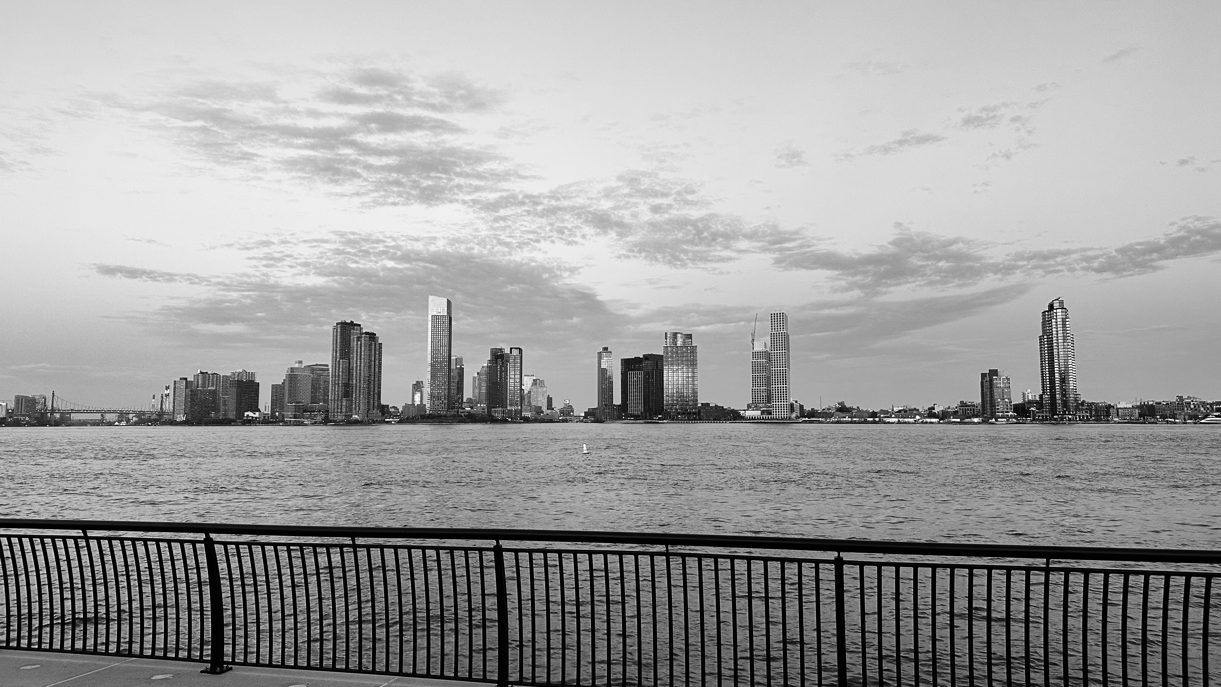 a black and white photo of the east river,high rise buildings and railings from the pier.